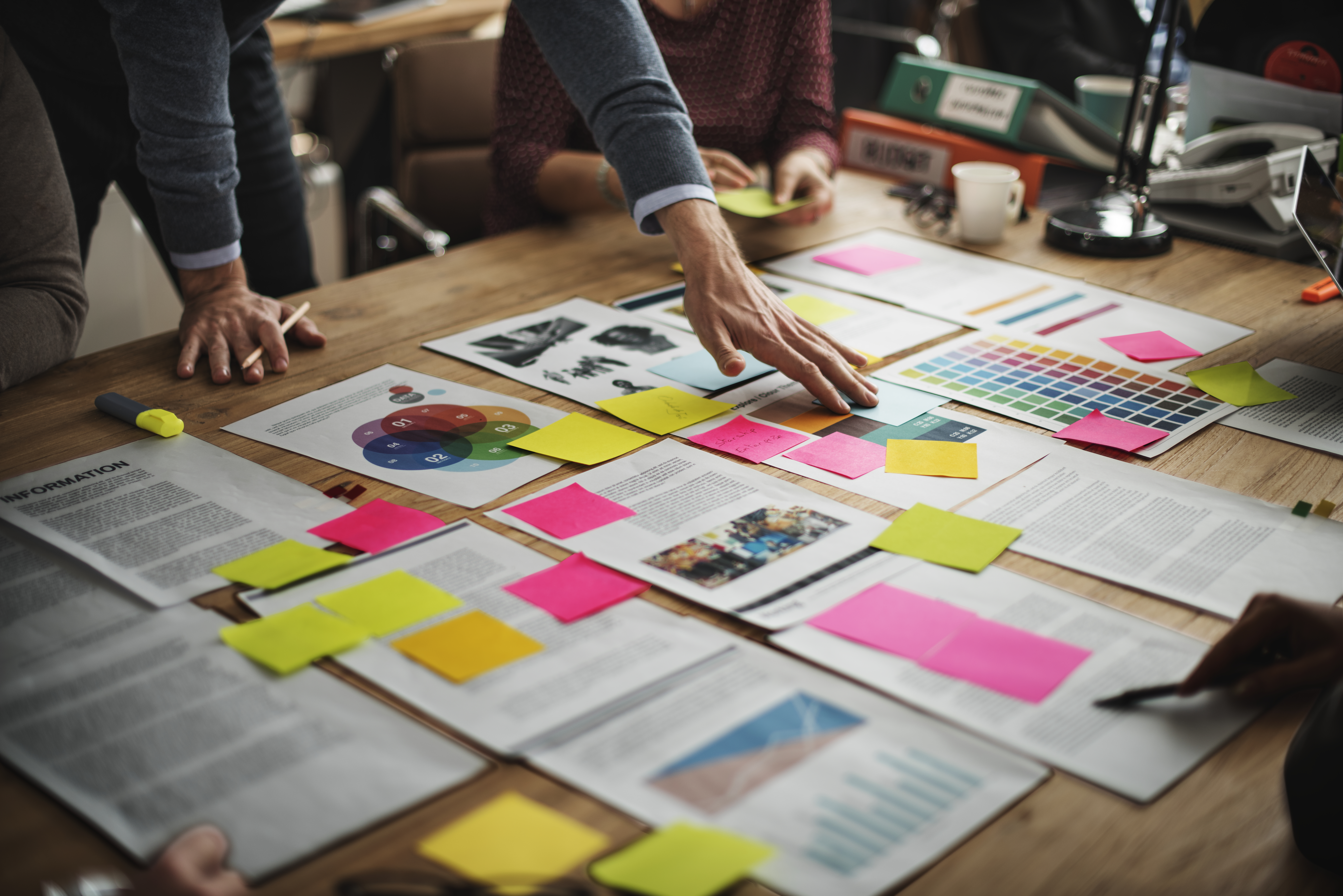 Mehrere Personen stehen um einen Tisch mit Papieren, Post-its in verschiedenen Farben und Statistiken; Hände zeigen auf Inhalte, symbolisiert Planung und Zusammenarbeit für Marketing-Ziele.