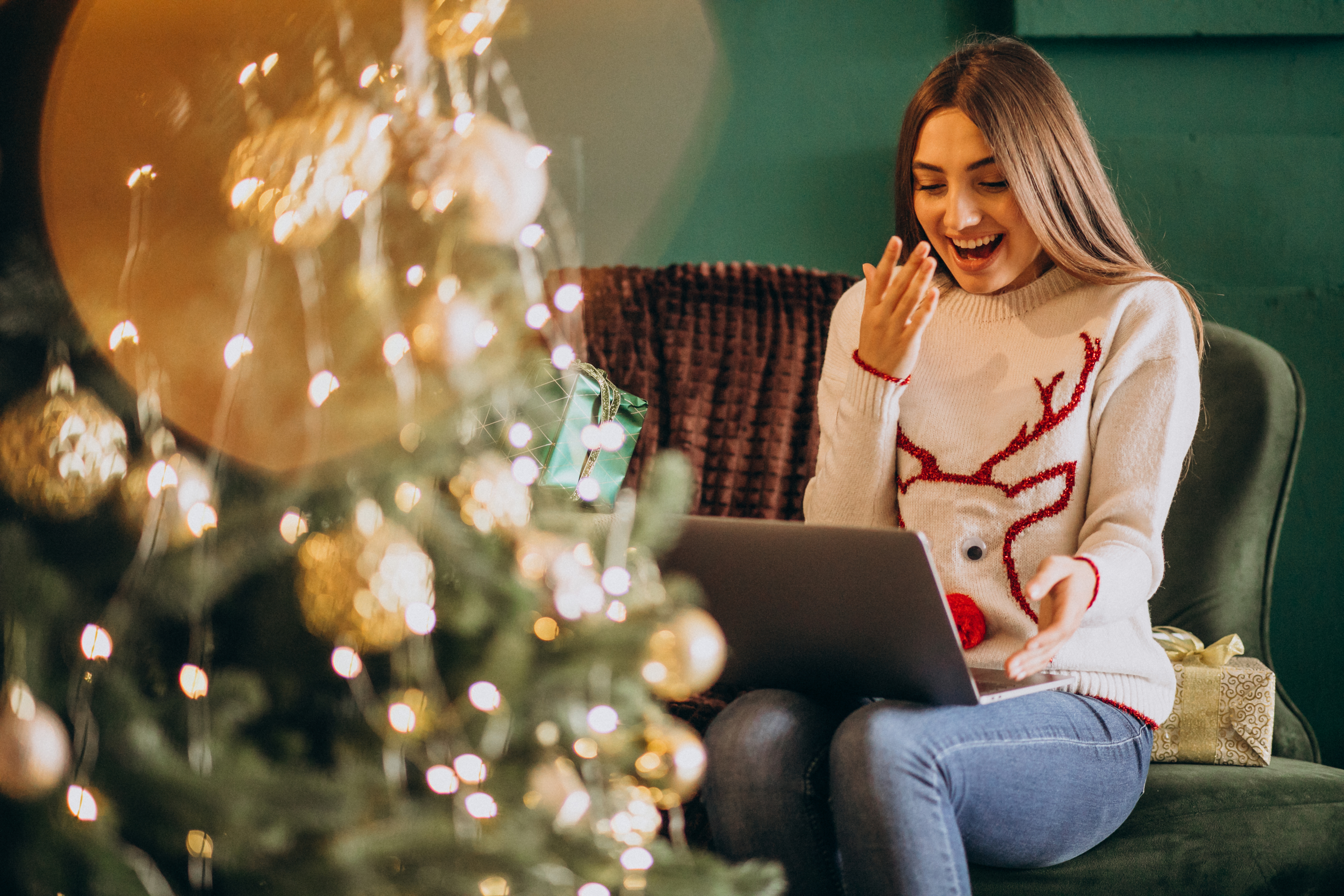 Frau in Weihnachtspullover sitzt mit geöffnetem Laptop auf dem Schoß, schaut glücklich und überrascht auf den Bildschirm; links im Bild ein geschmückter Weihnachtsbaum, symbolisiert digitale Weihnachtsmarketing-Aktivitäten.
